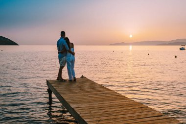 Girit Yunanistan 'da, birbirine aşık genç romantik çift gün doğumunda altın gökyüzü eşliğinde kumsaldaki ahşap iskelede oturup sarılıyor. Tatil ve seyahat konsepti. Deniz kenarında flört eden romantik genç çift