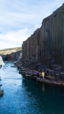 Ziyaretçiler, Studlagil Kanyonu 'nun etkileyici bazalt sütunlarına hayranlık duyuyor. Turkuaz sular, berrak bir gökyüzü altında dramatik bir manzara boyunca akıyor. İzlanda 'da görülmesi gereken bir doğa harikası.