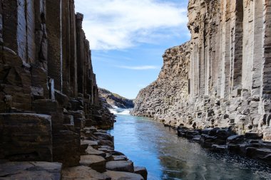 Studlagil Kanyonları 'nda bazalt sütunlara çarpıp heybetli bir şekilde yükselen sakin bir nehir. Bu nefes kesici İzlanda simgesi saf güzelliği berrak mavi gökyüzüne karşı sergiliyor..