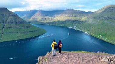 Faroe Adaları 'ndaki dramatik bir uçurumun tepesinde iki maceracı duruyor. Aşağıdaki yemyeşil tepelerin ve sakin suların büyüleyici panoramik manzarasının tadını çıkarıyorlar, alabalık çemberleriyle dolu. 