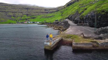 İki kişi Tjörk 'ün sakin sularında bir taş iskelede duruyor. Bereketli tepeler ve geleneksel Faroe Adaları evleri bu manzaranın güzelliğini temsil ediyor..