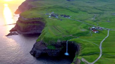 Güneş batarken Atlantik 'e dökülen Mullafossur Şelalesi' nin çarpıcı manzarası, Gasadalur, Faroe Adaları 'ndaki yemyeşil tepeler ve tuhaf köy yapılarıyla çevrili..