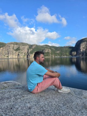 Norveç 'te sakin bir fiyorda bakan kayalık bir çıkıntıda huzur içinde oturan bir adam. Sakin sular görkemli uçurumları ve mavi gökyüzünü yansıtıyor. Doğayla iç içe olma ve bağlantı kurma davetkar anları..