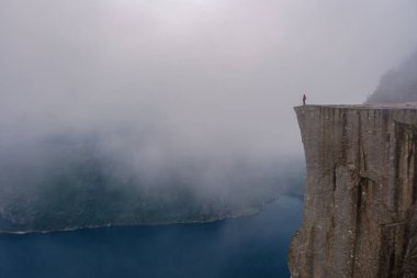 Norveç 'teki büyüleyici Lysefjord' un üzerinde, sisle kaplanmış dramatik uçurumlar görkemli bir şekilde yükseliyor. Yalnız bir yürüyüşçü Preikestolen 'in kenarında duruyor, doğanın huzurlu güzelliğini kucaklıyor..