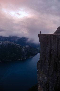 Preikestolen Cliff 'in kenarında duran yalnız bir figür aşağıdaki büyüleyici Lysefjorden' a bakıyor. Günbatımının ılık renkleri dramatik manzarayı aydınlatır, doğanın güzelliğini ortaya çıkarır..