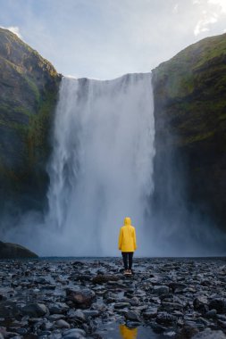 İzlanda 'daki Skogafoss şelalesinin nefes kesici manzarası. Sarı yağmurluklu bir şahıs, parçalı bulutlu bir gökyüzünün altındaki çağlayan sulara bakıyor..