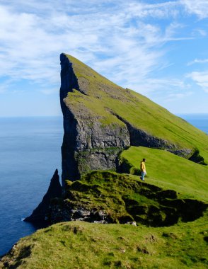Tek başına bir gezgin, Faroe Adaları 'ndaki keskin, yüksek bir uçurumun kenarında duruyor. Etrafı yemyeşil ve okyanusun panoramik manzaralarıyla çevrili. Açık gökyüzü manzaralı güzelliği arttırır.