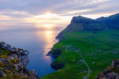 Altın güneş ışığı Faroe Adaları 'nın dramatik uçurumlarına sıcak bir parıltı saçarak aşağıdaki sakin sulardan yansıyor. Gasadalur köyü yakınlarında Mulafossur şelalesi