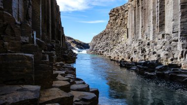 Studlagil Kanyonu, yukarıdaki açık mavi gökyüzünü yansıtan sakin bir nehirden yükselen şaşırtıcı bazalt sütun oluşumlarını sergiliyor. Bu doğa harikası İzlanda 'daki macera arayanlar için bir ziyaretti.
