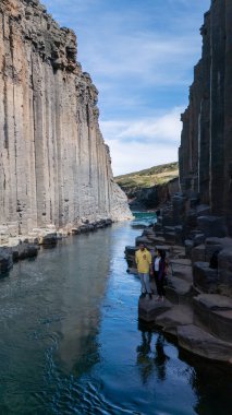 Ziyaretçiler, İzlanda 'nın yüksek bazalt kayalıklarıyla çevrili Studlagil Kanyonu' nun sakin sularının yanında duruyorlar..
