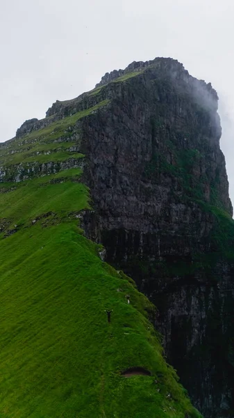 Faroe Adaları 'nın canlı yeşil tepelerinde bulutlu bir gökyüzünün altında çarpıcı bir uçurum yükselir. Bulutlar yavaşça kayalık tepenin etrafında sürükleniyor ve bu eşsiz manzaranın özünü yakalıyor..