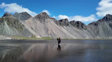 Bir çift adam ve kadın Stokksnes sahilinin sakin siyah kumlarında parlak mavi gökyüzünün altındaki görkemli Vestrahorn dağını yansıtıyorlar. Doğanın saf güzelliği cezbeder.