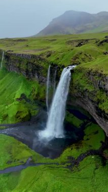 Seljalandsfoss şelalesinin yeşil İzlanda manzarasına çarpıcı bir şekilde düşüşünü deneyimleyin..