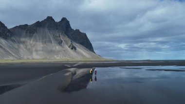 İki maceraperest, arka planda nefes kesen dağlarla İzlanda 'daki uzak bir siyah kumsal sahilinde geziniyor. Gökyüzü bulutlarla kaplı, sakin ve huzurlu bir atmosfer yaratıyor..
