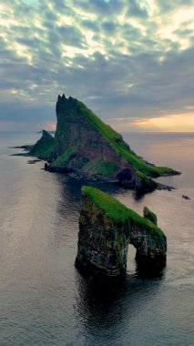 Faroe Adaları 'ndaki Drangarnir uçurumlarının gün batımında sakin suların üzerinde kule gibi uzanan nefes kesici güzelliğini deneyimleyin. Bu çarpıcı doğa harikası yeşil manzaraları ve engebeli uçurumları sergiliyor..