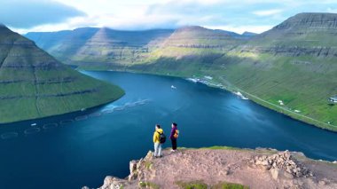 İki maceracının sakin sulara ve görkemli yeşil tepelere bakan kayalık bir uçurumun tepesinde durduğu çarpıcı Faroe Adaları 'na yolculuk. Klakkur zirvesinde bir çift kadın ve erkek.