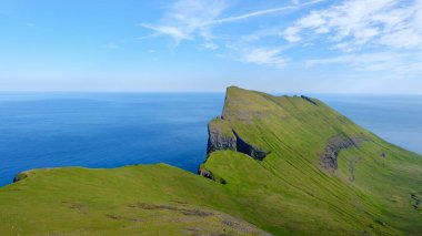 Gökyüzüne doğru yükselen yeşil kayalıklar, Faroe Adaları 'ndaki sakin denize bakıyor. Bu nefes kesen kıyı manzarası, açık mavi gökyüzünün altındaki bu uzak adaların eşsiz manzarasını gösteriyor..