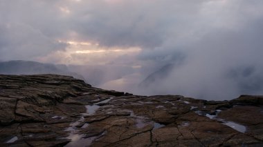 Preikestolen Cliff 'in dramatik manzaralarını keşfedin, sis engebeli araziyi kaplarken ve güneş dağların ardında batarken. Norveç 'te doğanın dingin güzelliğini tecrübe edin..