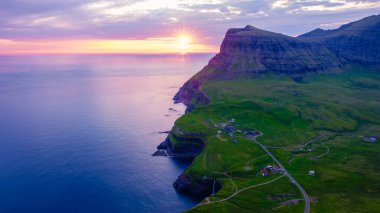 Güneş, Faroe Adaları 'nın dramatik uçurumlarının ardında batarken, aşağıdaki yemyeşil tepeleri ve sakin suları aydınlatan canlı renkler gökyüzünü dolduruyor. Gasadalur köyü yakınlarında Mulafossur şelalesi
