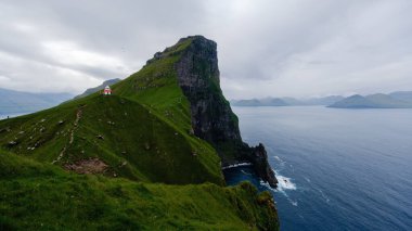 Dramatik bir uçuruma kurulmuş canlı kırmızı ve beyaz bir deniz feneri yeşil tepelerin ve karanlık okyanus dalgalarının önünde dimdik ayakta duruyor. Kallur Deniz Feneri, Kalsoy Adası