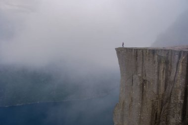 Norveç 'teki Preikestolen Cliff' in nefes kesen güzelliğini deneyimleyin. Aşağıdaki derin mavi sulara tepeden bakan yalnız bir figür mistik bir sis atmosferiyle kaplıdır..