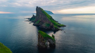 Faroe Adaları 'ndaki dingin mavi suların üzerindeki yemyeşil kuleyle kaplı dramatik Drangarnir uçurumları. Gün batımındaki ışık, bu uzak yerin doğal güzelliğini arttıran altın bir parıltı saçar..