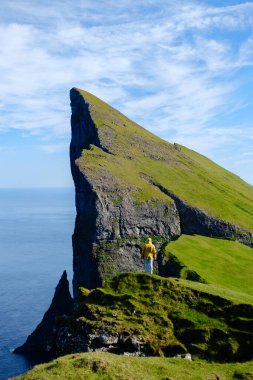 Faroe Adaları 'ndaki yemyeşil bir uçurumda duran bir gezgin dramatik Mylingur kaya oluşumlarına ve okyanusa bakıyor. Parlak mavi gökyüzü nefes kesen manzarayı güçlendirir..