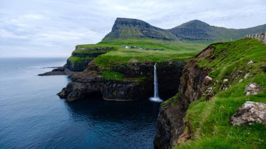 Görkemli uçurumlar Faroe Adaları 'ndaki berrak suların üzerinde dramatik bir şekilde yükselir. Gasadalur köyü yakınlarında Mulafossur şelalesi