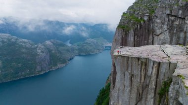Ziyaretçiler, Norveç 'in dramatik fiyort manzarasına hayret ederek Preikestolen Cliff' in kenarında duruyorlar. Sakin mavi sular engebeli yeşil tepelerle tezat oluşturuyor. Bulutlu bir gökyüzü atmosfere ekleniyor.