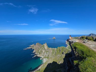 Bleik, Andoya, Norveç 'teki Matind Dağı' nın çarpıcı manzarasını görün. Yalnız bir yürüyüşçü kayalık kıyıda oturur, turkuaz sulara ve berrak mavi gökyüzünün altında dramatik uçurumlara hayret eder..