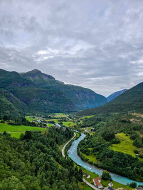 Görkemli dağlar, Fortun Norveç 'te canlı bir nehrin yemyeşil tarlalarda ve bulutlu bir gökyüzü altında büyüleyici evler arasında dolaştığı pitoresk bir vadi oluşturur. Doğa güzelliği burada gelişir..