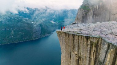 İki maceraperest yürüyüşçü, görkemli Lysefjord 'a tepeden bakan Preikestolen Cliff' in kenarında duruyor. Dramatik dağlar ve sisli gökyüzüyle çevrili bu ikonik nokta nefes kesici manzaralar sunuyor..