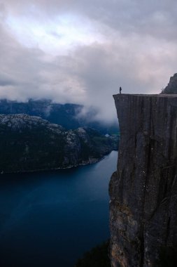 Preikestolen Cliff 'in kenarında duran yalnız bir figür çarpıcı Lysefjord' a bakıyor, dramatik dağlar ve bulutlarla çevrili, şafak vakti Norveç 'in dingin güzelliğini yakalıyor..