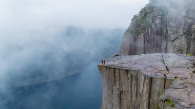 Maceracılar, nefes kesici Lysefjord 'a tepeden bakan Preikestolen Cliff' in kenarında dururlar. Sisler dramatik manzarayı sarar ve yolculukları boyunca ruhani bir atmosfer yaratır..