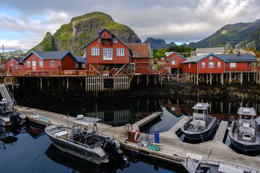  Bir Lofoten, Norveç, 13 Ağustos 2025, rıhtımdaki renkli kırmızı kulübeler, arka planda dağların parçalı bulutlu bir gökyüzünün altında belirmesiyle botlar huzur içinde kenetlendi,