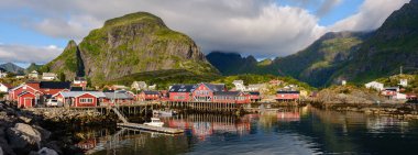 Canlı balıkçı kulübeleri A Lofoten, Norveç 'in sakin sularını yansıtır. Parçalı bulutlu bir gökyüzünün altında arkaplanda yükselen görkemli dağlar gün ışığında nefes kesen bir kıyı manzarası yaratıyor..