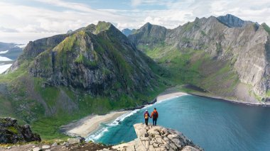 İki yürüyüşçü Ryten zirvesinden Kvalvika Sahili 'nin turkuaz sularına bakarak büyüleyici manzaraya hayran kaldılar. Bu unutulmaz an, Lofotens 'ın engebeli arazisinin ve yemyeşil toprağının güzelliğini yakalar..