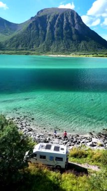 Norveç 'in Lofoten kentindeki bir fiyordun ışıldayan turkuaz sularının yanına park etmiş bir karavan manzarası. Bir çift, kapalı park etmiş bir karavanda yolculuk yapıyor.