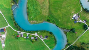Norveç 'in Stryn kentindeki yemyeşil tarlalar, açık gökyüzünün altındaki dolambaçlı turkuaz nehri çevrelemektedir. Tuhaf evler manzarayı süslüyor, huzur ve cazibeyle dolu pitoresk bir kırsal ortam yaratıyor..
