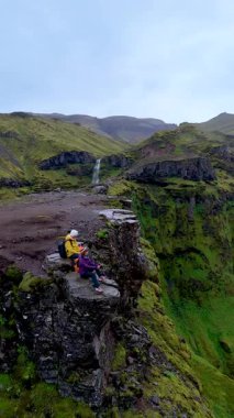 İzlanda 'daki Mulagljufur Kanyonu' nun büyüleyici manzaralarını keşfedin. Bir çift adam ve kadın bir kayanın üzerinde oturup şelaleye bakıyorlar.