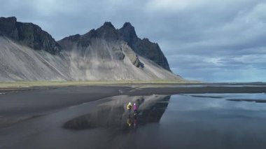 Maceracılar, karamsar bir gökyüzünün altında yükselen dağlarla çevrili İzlanda sahillerinin dramatik kara kumlarını aşarlar. Stoksnes Burnu ve Vestrahorn Dağı