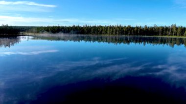 Finlandiya 'daki Hossa Ulusal Parkı' nda bir gölün dingin güzelliğini tecrübe edin. Kristal berrak sular mavi gökyüzünü ve yemyeşil ormanı yansıtıyor, sisli bir gündoğumunda doğaya sakin bir kaçış yaratıyor.