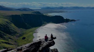 Bir çift, Norveç 'in Matind Andoy kentinin çarpıcı kıyı manzarasına bakarak kayalık bir çıkıntıda oturuyor. Kristal berrak su ve etrafındaki dağlar huzurlu bir atmosfer yaratır..