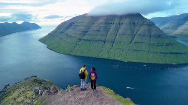 İki maceracı Faroe Adaları 'ndaki bir uçurumun tepesinde durup sakin sulara ve görkemli tepelere bakıyorlar. Klakkur zirvesinde bir çift kadın ve erkek.