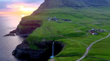 Mullafossur şelalesi, güneş batarken büyüleyici yeşil tepeler ve uzak kayalıklarla çevrili Gasadalur, Faroe Adaları 'ndaki okyanusa çarpıcı bir şekilde akmaktadır..