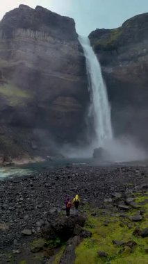 İki kaşif, muhteşem Haifoss şelalesinin kayalık kayalıklardan aşağı yuvarlanışına hayranlıkla bakıyor. Şelaleden sis yükselir ve etraflarındaki yemyeşil manzara büyüdükçe mistik bir atmosfer yaratır..