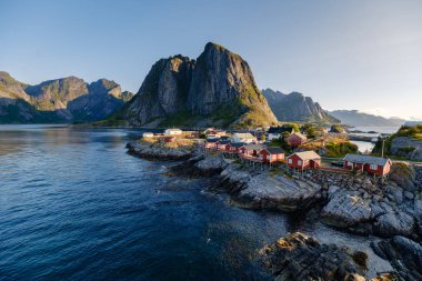 Hamnoy köyünde Norveç 'in Lofoten kentindeki çarpıcı dağ arkaplanlarına karşı geleneksel kırmızı balıkçı kulübeleri sergileniyor. Altın saat boyunca yakalanan sakin sular nefes kesici manzarayı yansıtıyor..