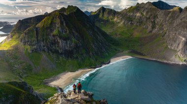 Yürüyüşçüler Ryten Dağı 'nın zirvesinden Kvalvika Sahili' ne bakan büyüleyici manzaranın keyfini çıkarıyorlar. Bereketli yeşillik, canlı bir gökyüzünün altında ışıl ışıl okyanusla güzel bir tezat oluşturuyor..