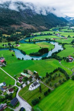 Norveç 'in Stryn kentinin çevresindeki yemyeşil alanlar boyunca uzanırken kıvrımlı bir nehir zarifçe vadiden akar. Görkemli dağlar bulutlu bir gökyüzünün altında çarpıcı bir zemin oluşturur..