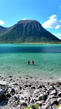 İki kişi, güneşli bir günde, parlak mavi gökyüzünün altında yükselen dağlarla çevrili Lofoten 'deki Norveç fiyortunun berrak turkuaz sularında ferahlatıcı bir yüzüşten zevk alır..